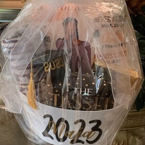 May include: A graduation gift basket with a white and gold tassel, a black and gold box with the text "The Tassel", a black graduation cap with the year "2023" in gold lettering, and a white gift bag with the text "2023" in gold lettering.