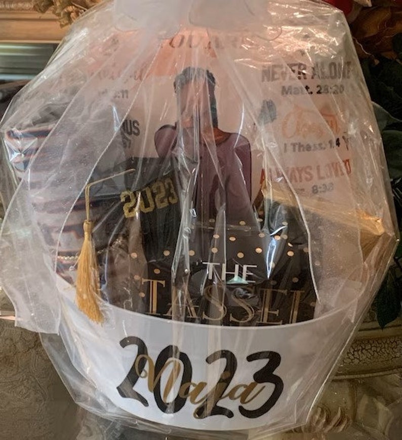 May include: A graduation gift basket with a white and gold tassel, a black and gold box with the text "The Tassel", a black graduation cap with the year "2023" in gold lettering, and a white gift bag with the text "2023" in gold lettering.