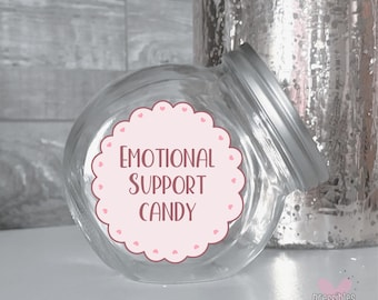 May include: A clear glass jar with a silver lid, labeled "EMOTIONAL SUPPORT CANDY" on a pink scalloped sticker with heart accents. The jar is slightly tilted, and a decorative silver vase is in the background.