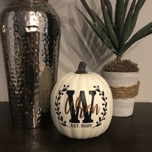 May include: A white pumpkin with a black and brown design. The design includes a black letter "W" inside a wreath, the name "Wren" in brown script, and the text "EST. 2023". A decorative fall-themed item.
