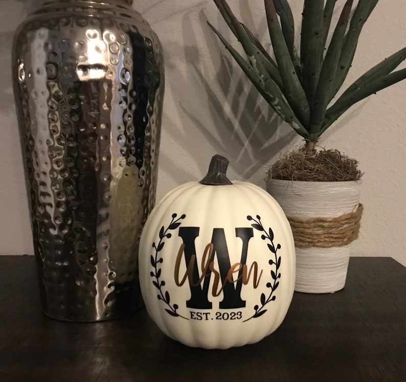 May include: A white pumpkin with a black and brown design. The design includes a black letter "W" inside a wreath, the name "Wren" in brown script, and the text "EST. 2023". A decorative fall-themed item.