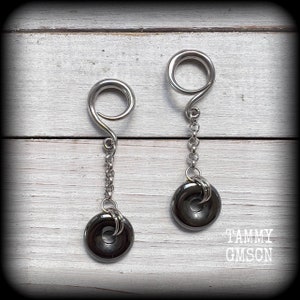 May include: A pair of silver-toned earrings with a spiral top design, connected by a chain to a dark gray, circular pendant. The earrings are displayed on a white wooden surface.