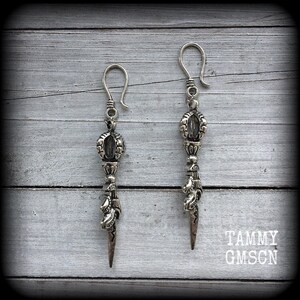 May include: A pair of silver-toned dangle earrings. Each earring features a hook, ornate central design, and a pointed bottom. The earrings are displayed on a light gray wooden surface. The text "TAMMY GMSCN" is visible in the lower right corner.