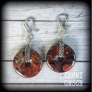 May include: A pair of handcrafted earrings featuring circular, reddish-brown stone pendants with silver-tone chain accents. The earrings have silver-tone ear wires and a unique, marbled appearance. The image includes the text "TAMMY GMSCN".