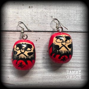 May include: A pair of red wooden earrings with a black and white painted face and the Japanese character for "luck" on the front. The earrings are hanging from silver hooks.