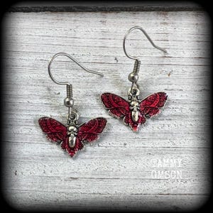 May include: A pair of dangle earrings featuring a moth-shaped charm with a skull design at the center. The moth wings are colored in a deep red with intricate black filigree patterns. The earrings have silver-toned metal hooks and hardware.