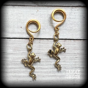 May include: A pair of gold-toned metal ear gauges with frog charms hanging from them. The gauges are circular and have a smooth finish. The frogs are detailed and have a realistic appearance.