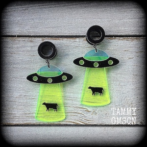 May include: A pair of black acrylic earrings featuring a UFO design with a neon green beam and a black cow silhouette. The UFO has a black disc with green lights and a blue and green top. The earrings have black plugs.