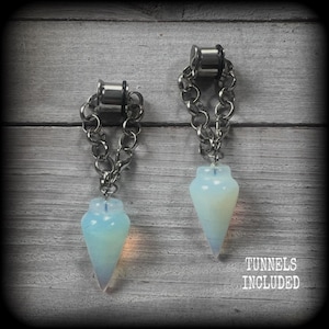 May include: A pair of silver-toned metal ear tunnels with attached silver-colored chains and dangling, cone-shaped, translucent white opal-like stone pendants. The text TUNNELS INCLUDED is visible in the bottom right corner of the image.