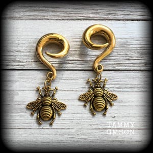 May include: A pair of gold-toned ear gauges featuring a spiral-shaped top and a dangling bee charm. Each piece is crafted with a metallic finish, showcasing a detailed insect design with textured wings and a striped body hanging from a small jump ring.