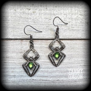 May include: A pair of dark gray spider earrings with green gemstone accents. The spider design features a geometric body and legs, with a teardrop-shaped green gem at the center. The earrings hang from a simple hook.