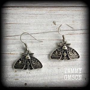 May include: A pair of silver-toned metal earrings featuring a moth design with intricate wing patterns, including crescent moon and circular motifs. The earrings hang from simple silver-toned hooks. The text TAMMY GMSCN is visible in the corner.