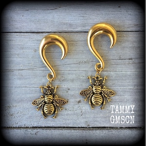 Peut inclure: Une paire d'écarteurs d'oreilles dorés en forme de crochet incurvé avec une breloque abeille suspendue à chaque pièce. Les breloques en métal présentent des ailes et des corps détaillés. Le texte TAMMY GMSCN est visible en bas à droite.