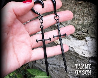 May include: A pair of black ear gauges featuring a curved hook design attached to a dangling sword pendant. The sword charms are detailed with a crossguard and a pointed blade. The text TAMMY GMSCN is visible in the lower right corner.