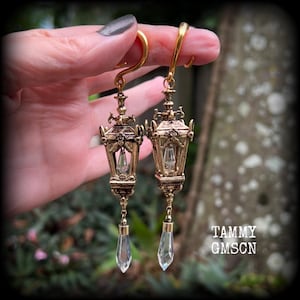 May include: Ornate gold-colored earrings in the shape of miniature lanterns. Each earring features a clear, teardrop-shaped crystal pendant. The earrings are held up by a gold hook. The earrings have intricate detailing and a vintage aesthetic.