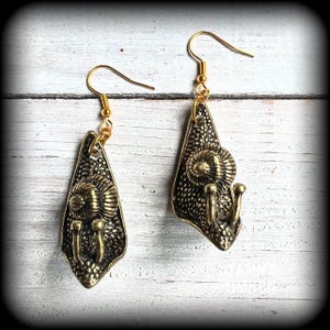 May include: A pair of gold-colored earrings. Each earring features a textured, teardrop-shaped pendant with a central spiral design and three small, vertical protrusions. The earrings have a hook closure.