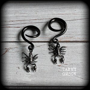 May include: A pair of black spiral earrings with a scorpion charm. The earrings have a curved, swirled design at the top and a detailed scorpion pendant hanging below. The earrings are displayed on a light gray wooden surface.