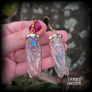 May include: A pair of cicada-shaped earrings featuring translucent, iridescent wings with a subtle blue and purple sheen. The insect bodies are encased in a clear material with gold-toned metal edges and matching gold-toned ear hooks for hanging.