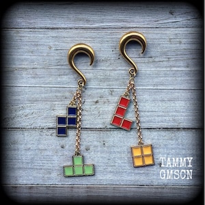May include: A pair of gold-colored earrings with a chain and colorful Tetris block charms. One earring features blue and green blocks, while the other has red and yellow blocks. The earrings are displayed on a light blue wooden surface.