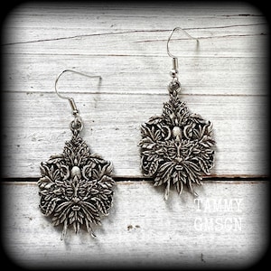 May include: A pair of silver-toned earrings featuring a Green Man design. Each earring has a detailed face surrounded by leaves and foliage, with a hook for pierced ears. The earrings are displayed on a white wooden surface.