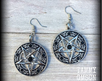 May include: A pair of silver-toned circular drop earrings featuring a central five-pointed star design enclosed within a ring. The metal surface has dark recessed detailing and small black circular accents. Each earring hangs from a simple hook wire.