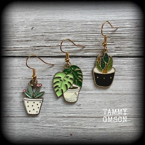 May include: Three pairs of gold-coloured dangle earrings with plant designs. One pair features a succulent in a white pot with black dots, another has a monstera leaf design, and the third has a succulent with pink flowers. The earrings are set against a light wood grain background.