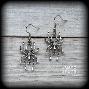 May include: A pair of silver-toned spider earrings. Each earring features a detailed spider design with a small, round, white bead at the center of the spider's body. The earrings hang from a simple hook. The background is a light-colored, weathered wooden surface.