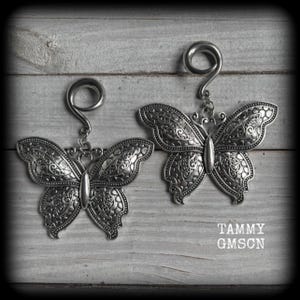 May include: Two silver-toned butterfly-shaped earrings with intricate etched details. Each earring features a decorative butterfly pendant suspended from a curved hook. The earrings are displayed on a weathered, gray wooden surface. The text "TAMMY GMSCN" is visible.