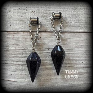 May include: A pair of dark blue plugs with a chain and a faceted, teardrop-shaped pendant. The plugs are made of silver-colored metal. The pendants have a speckled appearance. The image is set against a weathered wood background.