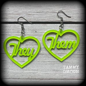 May include: A pair of neon green heart-shaped earrings. Each earring features the word "They" and "Them" in a cursive font. The earrings are suspended from silver-coloured ear wires. The background is a light grey wood grain.
