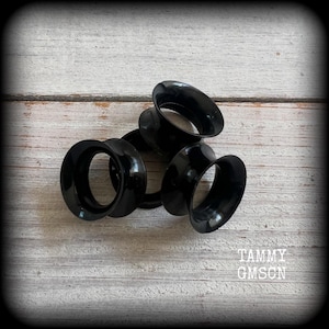 May include: Several black ear gauges are arranged on a light-colored, weathered wooden surface. The gauges have a smooth, glossy finish and a flared design. The text "TAMMY GMSON" is visible in the lower right corner.