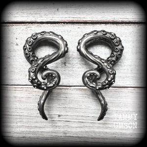 May include: A pair of silver-toned metal ear gauges shaped like octopus tentacles. The gauges have a textured surface and are designed to be worn in stretched earlobes.