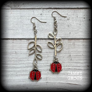 May include: A pair of silver-toned dangle earrings. Each earring features a leaf design and a red and black ladybug charm. The earrings are displayed on a white wooden surface. The text "TAMMY GMSON" is visible.