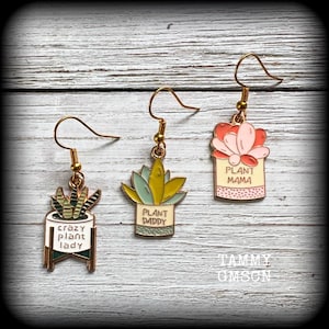 Op de afbeelding: Drie paar goudkleurige oorhangers met plantthema's. Eén paar zegt "crazy plant lady", een ander zegt "plant daddy" en de laatste zegt "plant mama". Elke oorbel heeft een ander vetplantontwerp.