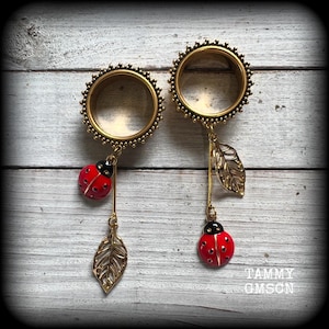 May include: A pair of gold-colored ear gauges with a decorative edge. Each gauge has a dangling charm featuring a red ladybug with black spots and a gold-colored leaf. The gauges are displayed on a light wood-grain surface.