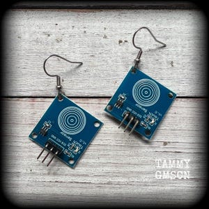 Op de afbeelding: Een paar blauwe touch-sensor oorbellen met zilveren haakjes. De sensoren hebben de tekst "Touch Sensor v1.0" en "SIG UCC GND" erop gedrukt.