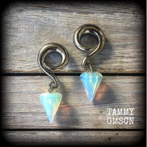 May include: A pair of silver ear gauges with opal teardrop charms. The gauges are shaped like a spiral and the charms are a light blue colour.