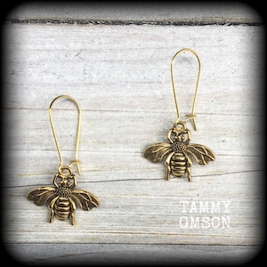 May include: A pair of gold-toned dangle earrings featuring a bee charm. The bee charm has a detailed design with wings, legs, and a segmented body.