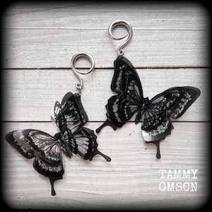 Peut inclure: Une paire de boucles d'oreilles papillon noires avec des détails argentés. Les ailes de papillon présentent des motifs détaillés et sont suspendues à des crochets argentés. Les boucles d'oreilles sont présentées sur une surface en bois clair. Le texte "TAMMY GMSCN" est visible.