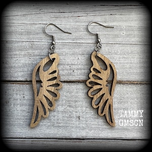 May include: A pair of wooden wing-shaped earrings. The earrings are light brown with intricate cut-out designs. Each earring has a silver-colored hook. The earrings are displayed on a light gray wooden surface. The text "TAMMY OMSCN" is in the bottom right corner.