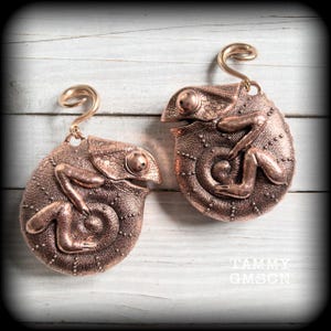 Peut inclure: Une paire de boucles d'oreilles couleur cuivre en forme de caméléons enroulés avec une peau texturée et des crochets en spirale. La surface métallique présente des motifs d'écailles et des membres en relief. Le texte TAMMY GMSCN est visible.