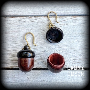 May include: Acorn-shaped earrings with gold-colored hooks. The earrings are made of dark brown and reddish-brown wood, with a black cap. One earring is assembled, and the other is disassembled, showing the inside of the acorn.