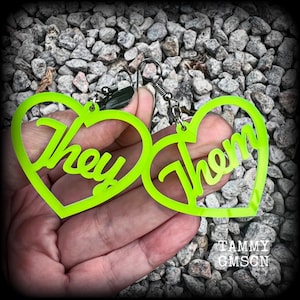 May include: Bright green heart-shaped earrings featuring the words "They" and "Them". The earrings are held up by a hand against a backdrop of small grey stones. The earrings have silver-coloured hooks.