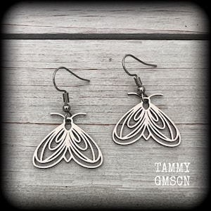 May include: A pair of silver-colored moth-shaped earrings. The earrings have a detailed, openwork design with multiple layers, and are suspended from simple ear wires. The earrings are displayed on a light-colored wooden surface. The text "TAMMY GMSCN" is visible.