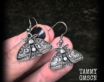 May include: A pair of silver-toned moth-shaped drop earrings featuring intricate wing patterns with crescent moon and circular motifs. The earrings hang from simple metal hooks. The text TAMMY GMSCN is printed in the bottom right corner.