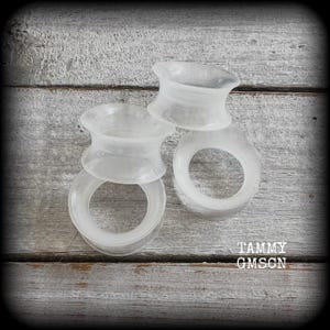 May include: Four translucent, white silicone ear gauges are stacked on a weathered, gray wooden surface. The gauges have a flared design, with a wider outer rim and a narrower center. The text "TAMMY GMSCN" is visible in the lower right corner.