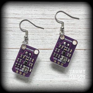 Op de afbeelding: Een paar unieke oorbellen met een paars printplaatontwerp. Elke oorbel heeft een zilveren haak en een rechthoekige hanger met de tekst "MAX4466" en andere elektronische componenten. Een creatief accessoire.