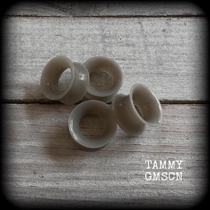 May include: Five gray ear gauges are arranged on a weathered, light gray wooden surface. The gauges are circular with a flared design. The text "TAMMY GMSCN" is visible in the lower right corner.