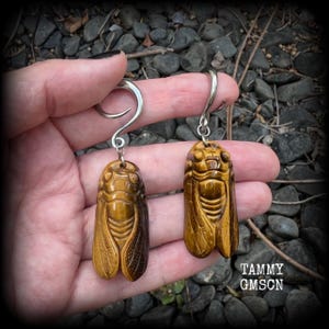 May include: A pair of ear weights featuring carved tiger eye stone cicada pendants suspended from silver-toned metal spiral hooks. The golden-brown, chatoyant stone insects display detailed wing and body segments, creating a naturalistic appearance.