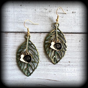 May include: A pair of dangle earrings featuring a cutout bronze-toned metal leaf charm suspended from a gold-toned hook. Attached to the center of each leaf is a small, white and black enamel snail charm. The text TAMMY GMSON is visible.